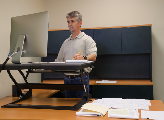 A man is working at a standup desk in an office where he works because standing is healthier than sitting all day. Live healthy, dont sit all day.